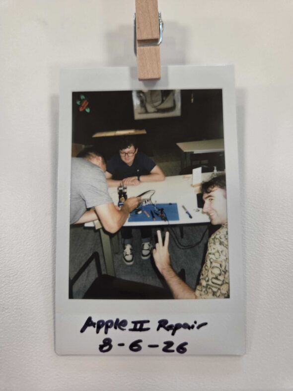 Polaroid photo of 3 men, one using a soldering iron to desolder the circuit board. Text says "Apple II Repair 8-6-26".