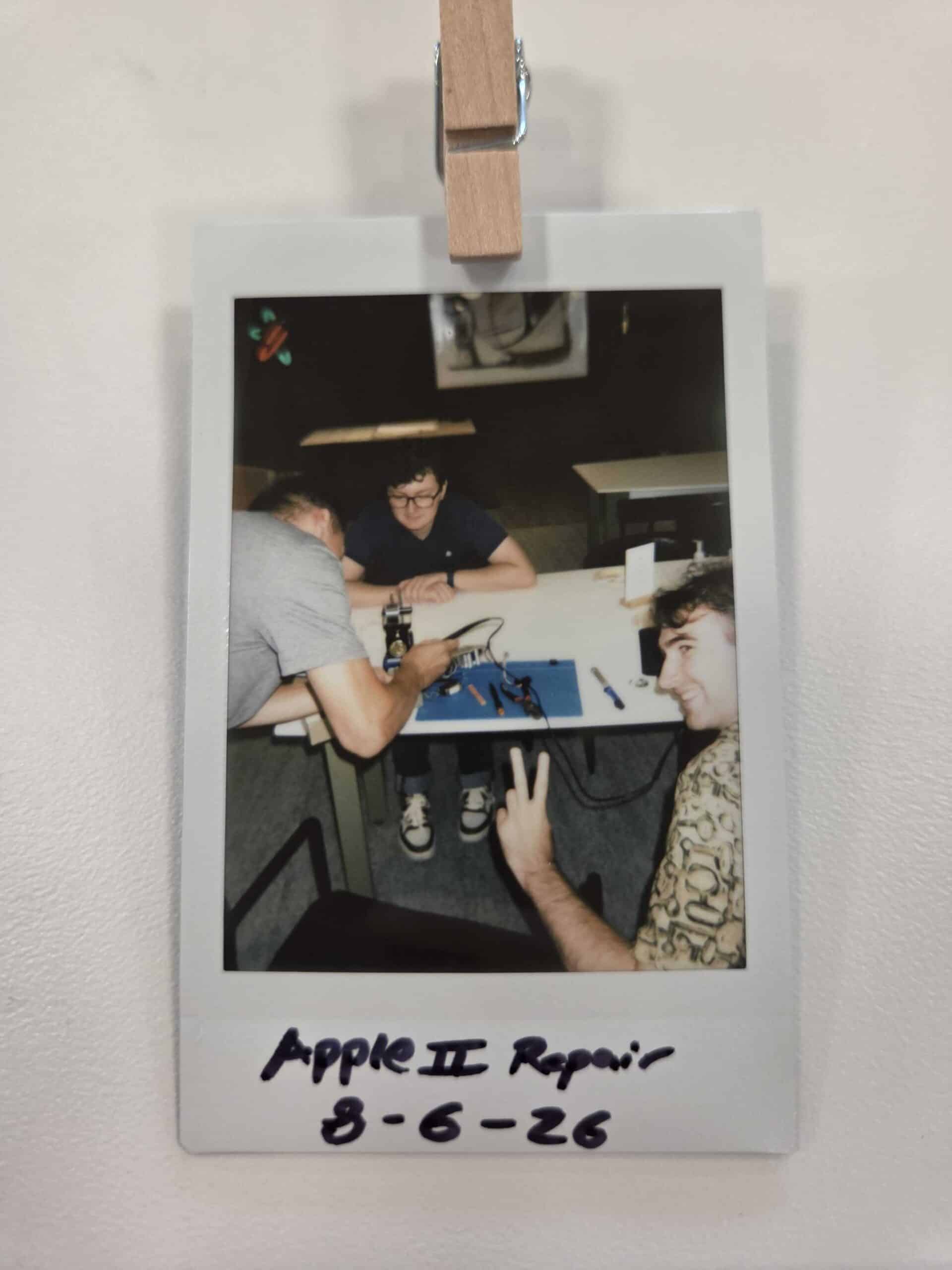 Polaroid photo of 3 men, one using a soldering iron to desolder the circuit board. Text says "Apple II Repair 8-6-26".