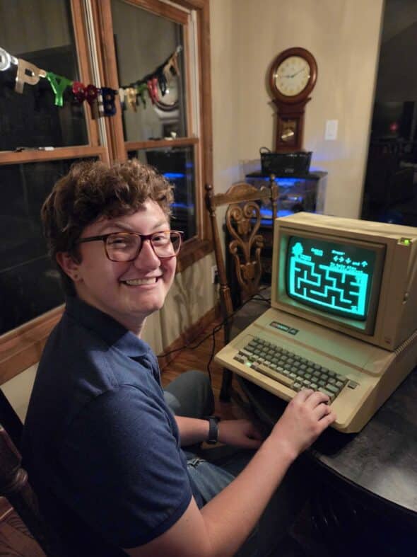 Jay, white male with curly brown hair and glasses, sits at the table smiling at the camera while his hand rests on the Apple IIe keyboard.