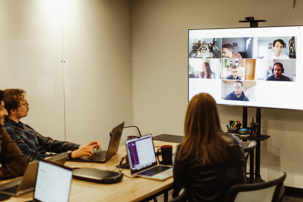 Atomic Object employees sitting around a table talking with remote employees on a screen. Post title: Quality Can’t Be Tested In — It Has to Be Built In