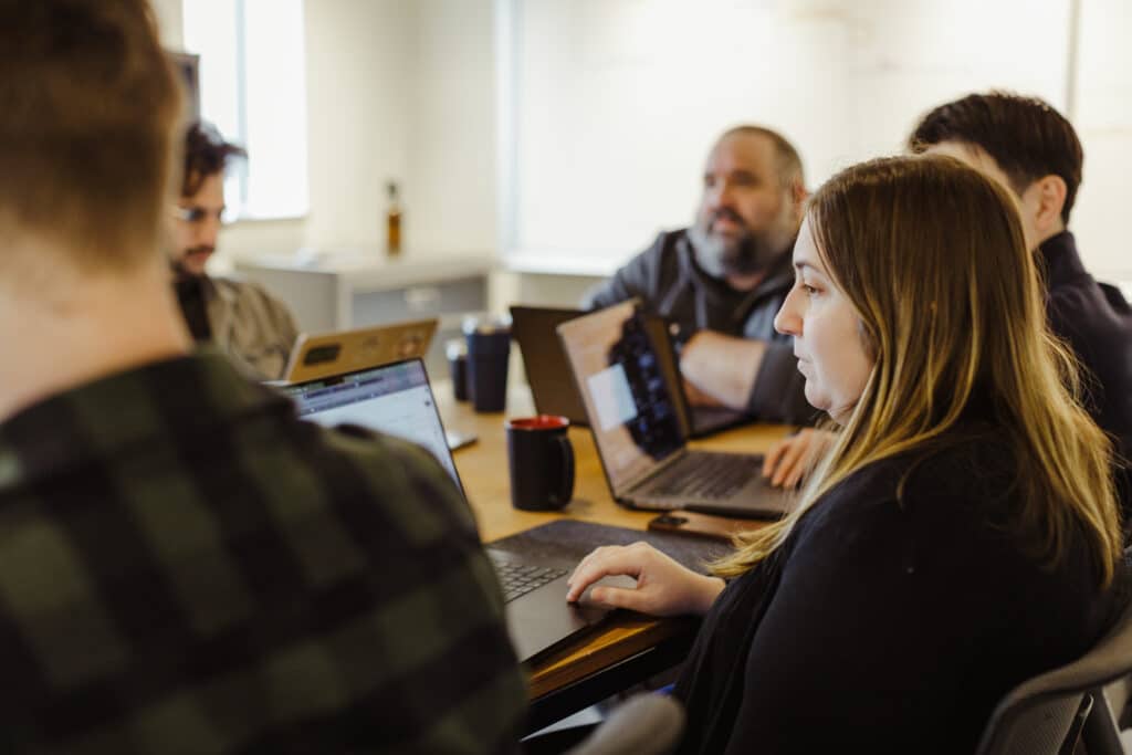 Atomic Object employees sitting at a table together in a team meeting. Post title: Steward, Strategist, Shield: The Essential Role of a Delivery Lead