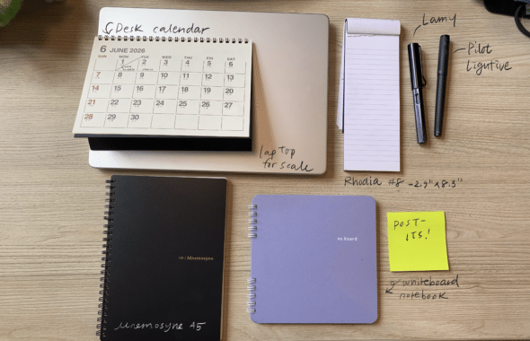Desktop calendar, long skinny notepad, black notebook, purple notebook, two fountain pens and a sticky note pad on a desk.
