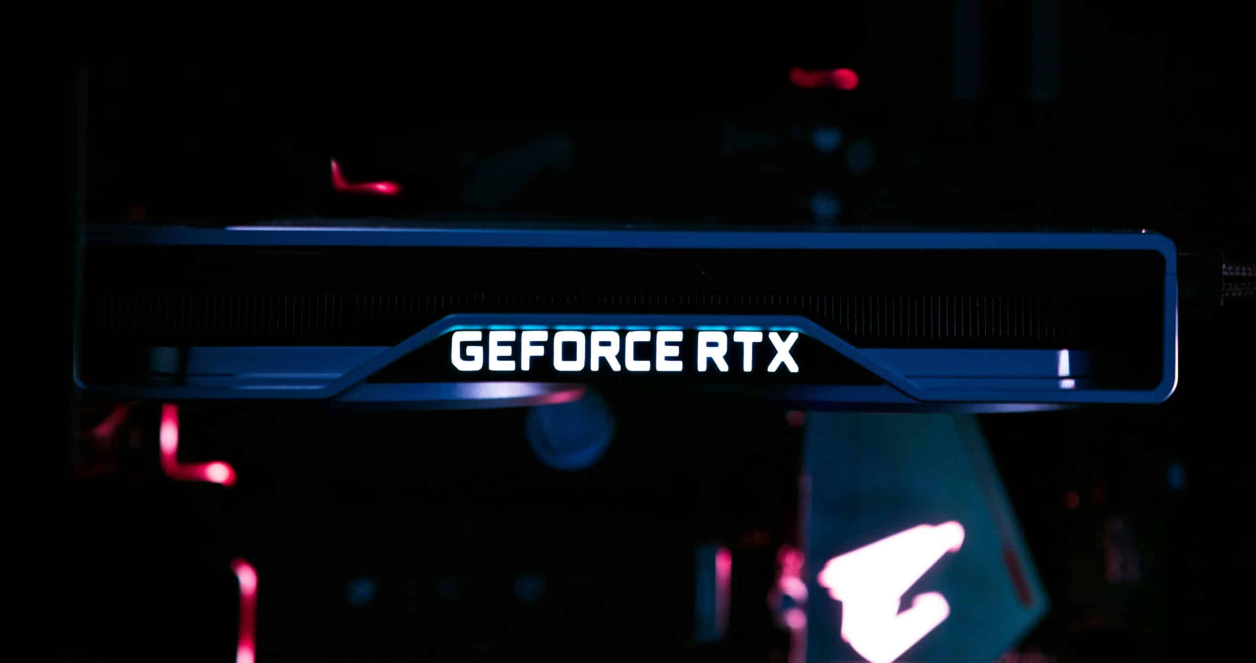 A close-up shot of an illuminated NVIDIA GeForce RTX graphics card installed in a desktop computer. The "GEFORCE RTX" branding is prominently lit in white on the side of the card, while the background is dark with subtle pink and blue lighting reflecting from other components inside the PC case.