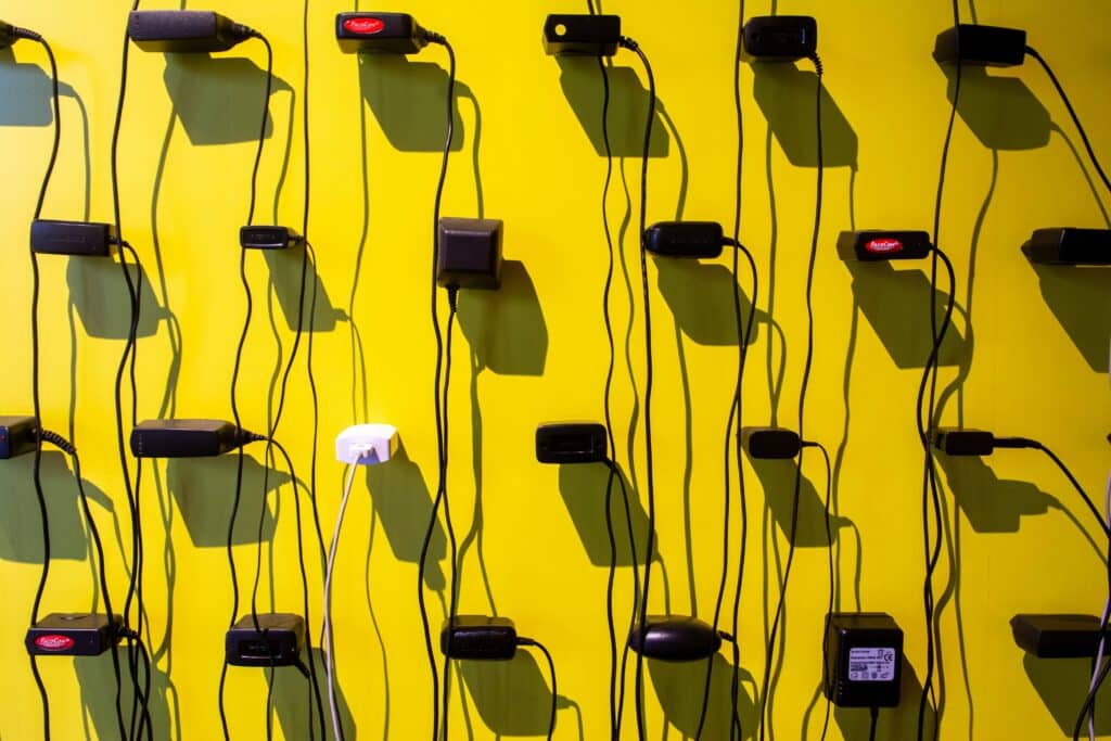 A wall of power adapters and tangled black cords plugged into outlets against a bright yellow background illustrating the post "Your Smart Home Shouldn’t Need Its Manufacturer to Stay Alive"