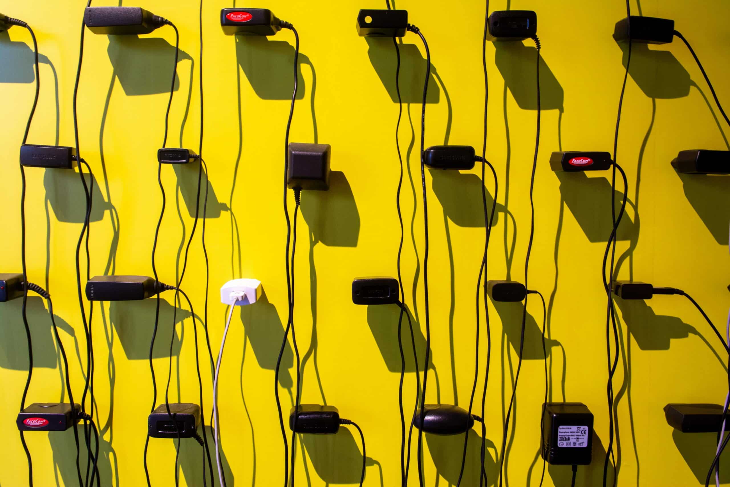 A wall of power adapters and tangled black cords plugged into outlets against a bright yellow background illustrating the post "Your Smart Home Shouldn’t Need Its Manufacturer to Stay Alive"