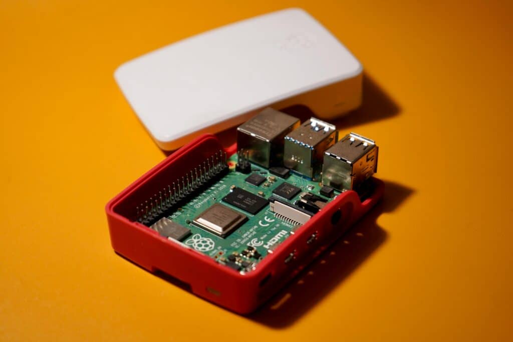 A Raspberry Pi single-board computer housed in a red case with the top cover removed, revealing the circuitry, ports, and GPIO pins. The top white cover with the Raspberry Pi logo is placed behind the board. The setup is resting on a solid yellow surface, and the lighting highlights the metal ports and green circuit board. Illustrating a piece called A Portable Pi Setup Guide