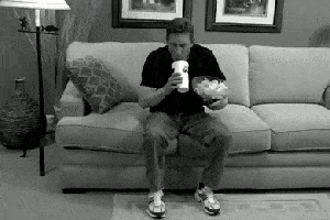 A hapless man on a couch spills popcorn and soda while trying to reach for the TV remote that’s in the hand that’s holding the popcorn.